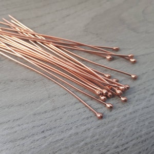 May include: A pile of rose gold head pins with rounded ends. The pins are arranged in a fan shape on a wooden surface.
