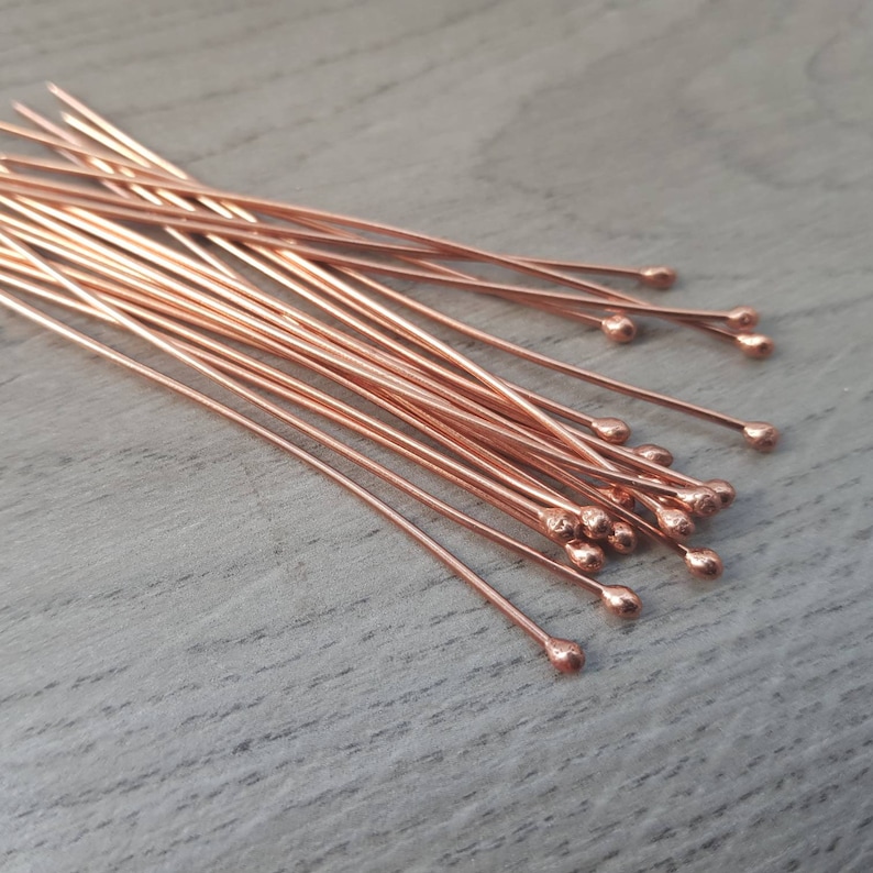 May include: A pile of rose gold head pins with rounded ends. The pins are arranged in a fan shape on a wooden surface.