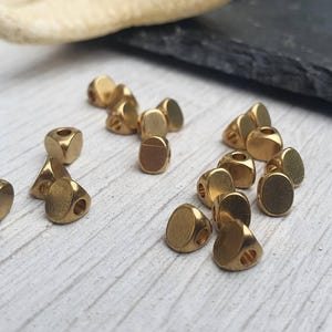 Peut inclure: Un ensemble de petites perles métalliques dorées. Les perles ont une forme géométrique aux bords arrondis et un trou au centre. Elles sont dispersées sur une surface en bois de couleur claire.