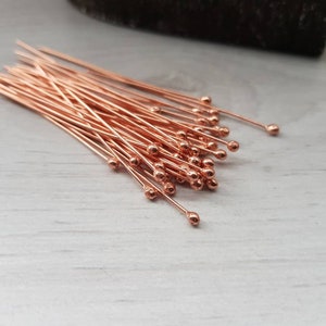May include: A bunch of rose gold head pins with round heads. The pins are arranged in a pile on a white surface.