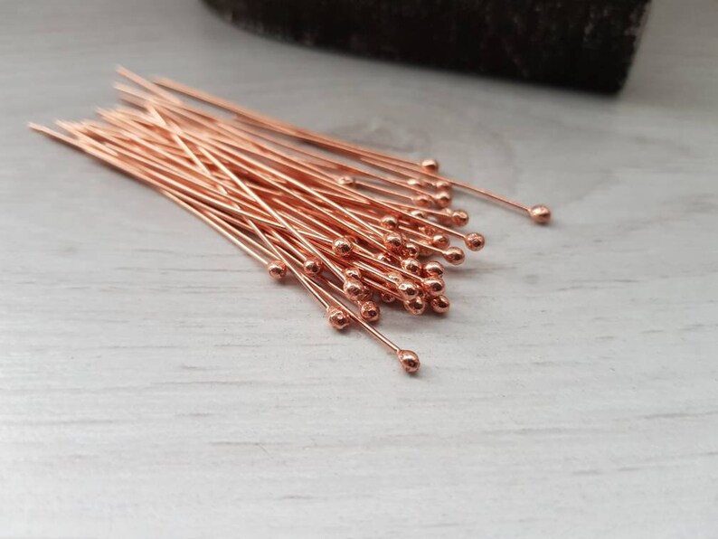 May include: A bunch of rose gold head pins with round heads. The pins are arranged in a pile on a white surface.