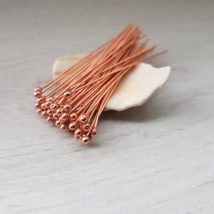 May include: A bunch of copper-colored head pins with round heads. The pins are arranged in a fan shape and are sitting on a white seashell.