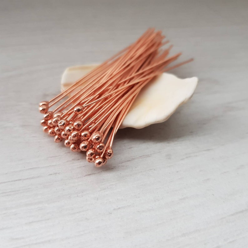 May include: A bunch of copper-colored head pins with round heads. The pins are arranged in a fan shape and are sitting on a white seashell.