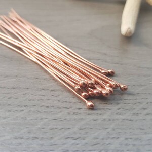 May include: A bundle of rose gold head pins with rounded ends. The pins are arranged in a fan shape on a wooden surface.