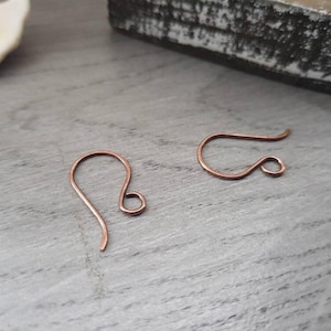 以下が含まれることがあります： 灰色の木製の表面に、2つの銅製のイヤリングフックが置かれています。フックは、細くて柔軟なワイヤーで作られており、シンプルでエレガントなデザインです。