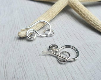 May include: Two silver-toned metal clasps with a swirl design. The clasps are on a white wooden surface.
