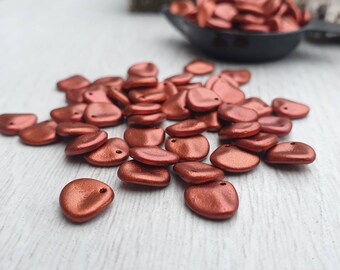 May include: A pile of small, copper-colored, metallic beads in the shape of a teardrop with a hole at the top. The beads are scattered on a white surface.