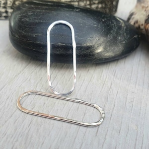 Peut inclure: Deux anneaux ovales en argent avec une texture martelée. Les anneaux sont posés sur une surface en bois clair.
