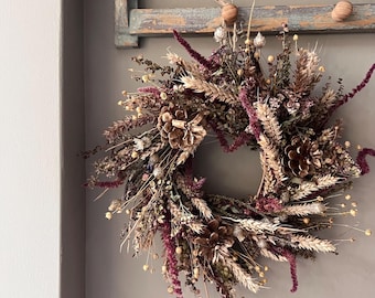 May include: A circular wreath composed of dried botanical elements including deep burgundy amaranth, golden wheat stalks, and brown pinecones. The arrangement features a variety of dried grasses, seed pods, and foliage, creating a textured display.
