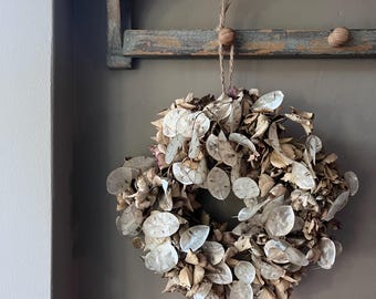 May include: A dried wreath made of natural materials, featuring a circular design with light brown and off-white elements. The wreath is suspended by a twine loop from a wooden shelf, creating a rustic, organic aesthetic. The wreath is a decorative item.