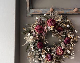 May include: A circular wreath composed of dried flowers and foliage in shades of deep burgundy, muted pink, cream, and earthy green. The arrangement features prominent dark red blooms interspersed with smaller white flowers and dried grasses.