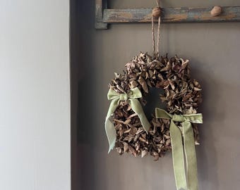 May include: A dried leaf wreath with two light green velvet bows. The wreath is hanging from a wooden rack with three wooden pegs. The background is a neutral brown color.