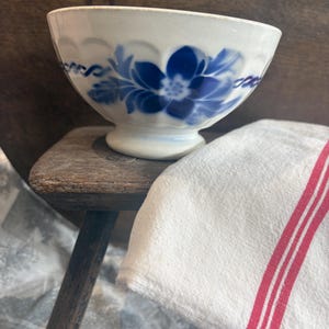 May include: A white ceramic bowl featuring a blue floral pattern and a scalloped rim. The bowl has a pedestal base and is positioned next to a white fabric cloth with two vertical red stripes, resting on a dark wooden surface.
