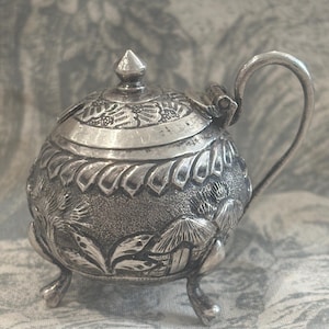 May include: A small, round silver-toned metal container with a hinged lid, a curved handle, and three short legs. The exterior features embossed details of palm trees and small huts, with a textured finish and a pointed finial on the lid.