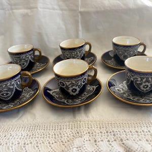 May include: A set of six small porcelain cups and matching saucers featuring a deep navy blue exterior with a white intricate lace-like floral pattern and gold trim along the rims and handles. The interior of each cup is a solid cream color.