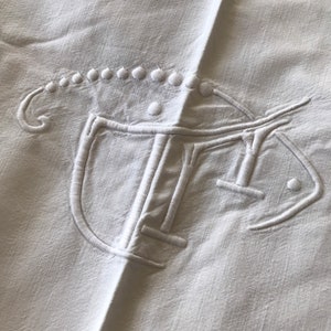 May include: White linen tablecloth featuring a large embroidered monogram in the centre. The monogram is a stylised letter "T" with a decorative border.