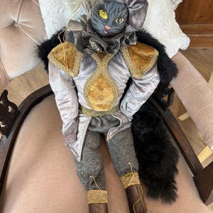 May include: A decorative doll featuring a cat head with a black and gray face, yellow eyes, and a feathered hat. The doll wears a gray velvet jacket with gold trim, a ruffled collar, gray trousers, and brown boots, accented by a black faux fur tail.