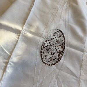 May include: Ivory-colored textile with an oval, openwork lace design. The lace features floral patterns and is set against a darker background. Vertical stitched lines and subtle folds add texture to the fabric.