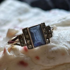 May include: A vintage ring with a rectangular, light blue gemstone set in a silver-toned metal frame. The ring features small, round accent stones and a delicate gold band. The design is Art Deco inspired.