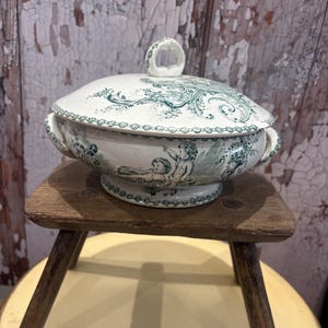 May include: A white ceramic tureen with a lid, featuring green transferware illustrations of cherubs and ornate scrollwork. The bowl has two side handles and a circular base, with a matching lid that includes a small, looped handle on top.