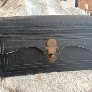 Op de afbeelding: Een vintage zwarte leren koffer met een messing sluiting. De rechthoekige koffer heeft een gebogen bovenkant en een klepsluiting. Het leer vertoont tekenen van slijtage, wat bijdraagt aan de antieke uitstraling. De sluiting is sierlijk.