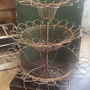 Könnte beinhalten: Ein dreistöckiger Obstkorb aus Draht mit einem dekorativen, gewellten Rand. Der Korb ist aus Metall und hat einen rustikalen, Vintage-Look.