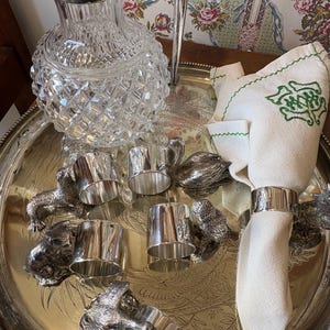 Puede incluir: Una bandeja plateada contiene una jarra de cristal, varios servilleteros plateados y una servilleta blanca doblada con bordados verdes. Los servilleteros presentan diseños de animales. La bandeja está sobre una superficie de madera.