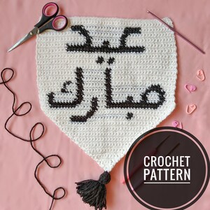 Op de afbeelding: Haakpatroon voor een wit en zwart gehaakt spandoek met de Arabische tekst "Eid Mubarak" in zwart. Het spandoek heeft een zwarte kwast aan de onderkant. De afbeelding toont ook een haaknaald en garen.