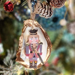 May include: A Christmas ornament featuring a wooden nutcracker soldier inside a gold-painted oyster shell. The nutcracker wears a black hat, red and purple uniform, and holds a sword. The ornament hangs from a natural twine string.
