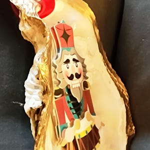 May include: A Christmas ornament featuring a nutcracker soldier design on an oyster shell. The ornament has a red ribbon for hanging. The shell is cream-colored with gold accents.