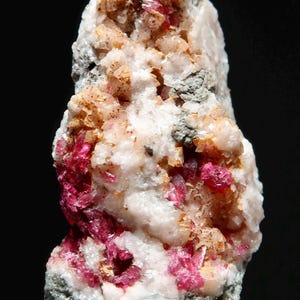 Puede incluir: Un cúmulo de cristales rosados y blancos sobre una roca gris. Los cristales son pequeños y tienen una textura rugosa. La roca mide aproximadamente 5 cm de alto.