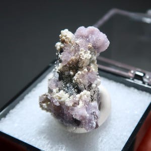 May include: A close-up of a mineral specimen featuring clusters of light purple, white, and dark grey formations. The mineral is presented on a white base within a black display case, showcasing its natural textures and colors.