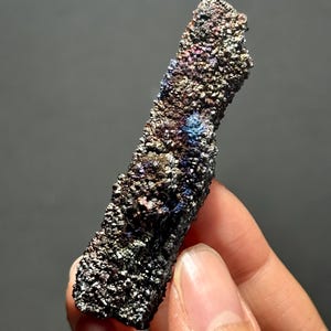 May include: A close-up of a mineral specimen with a rough, textured surface. The mineral displays a range of colors, including brown, purple, blue, and silver, with a metallic sheen. The specimen is held in a person's hand against a gray background.