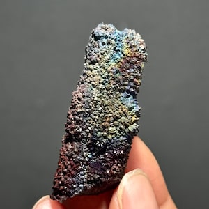 May include: A close-up of a mineral specimen with a rough, textured surface. The mineral displays a vibrant array of colors, including blues, greens, yellows, and reds, creating a rainbow effect. The specimen is held in a hand against a gray background.