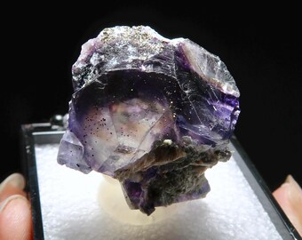 May include: A purple fluorite crystal with a light purple hue and a few small, dark inclusions. The crystal is sitting on a white surface in a black display box.