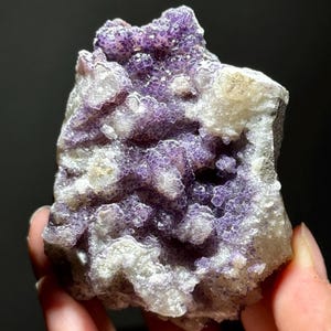 以下が含まれることがあります： 天然蛍石の鉱物標本をクローズアップ。石は紫と白の結晶構造が混ざっています。紫色の結晶は集まっており、質感のある表面を作り出しています。背景は暗く、鉱物の色を際立たせています。