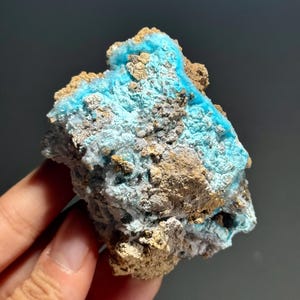 May include: A close-up of a mineral specimen featuring a vibrant turquoise-blue hue contrasted with earthy brown and gray tones. The rough, textured surface of the stone is visible, showcasing its natural formation and crystalline structure.