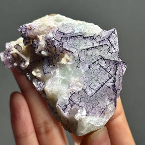 Può includere: Primo piano di un campione di minerale di fluorite. La pietra presenta un mix di colori, tra cui viola, bianco e sfumature di verde. Le sezioni viola hanno un motivo geometrico e la consistenza generale è cristallina.