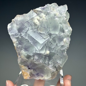 May include: A large, clear, purple fluorite crystal with a complex, geometric structure. The crystal is on a clear display stand with a label that reads "QR Fluorite, Dachang Mine, Qinglong, China."