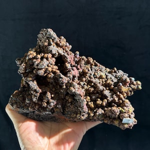 Peut inclure: Un spécimen minéral brut présentant une formation botryoïdale en grappe avec une texture sombre, métallique et brun terreux. La surface est couverte de nombreux petits nodules arrondis ressemblant à des cailloux avec des reflets bronze et cuivre.