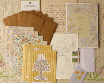 May include: A collection of stationery items, including cards, envelopes, and notepaper. The cards feature floral and cake designs, with a color palette of yellow, purple, and green. Some items have handwritten text. The stationery is arranged on a textured surface.