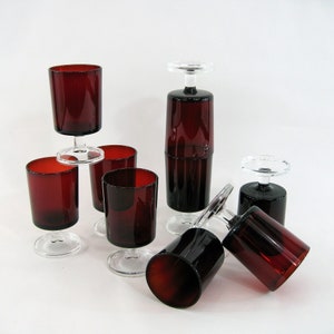 Può includere: Set di sette bicchieri da vino in vetro rosso con steli trasparenti. I bicchieri sono impilati e disposti su una superficie bianca.