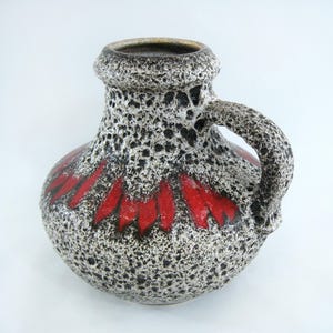 Peut inclure: Un vase en céramique avec une surface texturée et mouchetée dans des tons de gris et de noir. Le vase a un corps large et arrondi et une seule poignée. Une bande de glaçure rouge et noire décore la partie inférieure du vase.