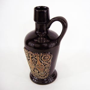 Può includere: Brocca in ceramica marrone scuro con manico e rilievo decorativo di uccelli e fiori. La brocca ha una finitura lucida e una base ampia. Il rilievo è di colore marrone chiaro.