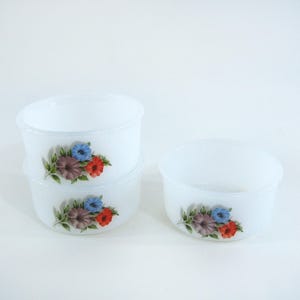 Könnte beinhalten: Set aus vier weißen Keramikschalen mit Blumendesigns. Die Schalen zeigen eine Gruppe von drei Blumen in Blautönen, Lila und Rot.