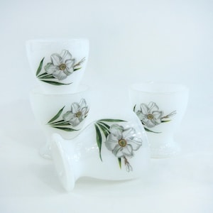 Könnte beinhalten: Set aus vier weißen gläsernen Eierbechern mit Blumenmuster. Jeder Becher zeigt eine weiße Blume mit grünen Blättern.