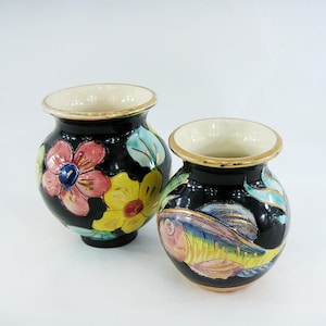 Könnte beinhalten: Zwei kleine schwarze Keramikvasen mit goldenen Verzierungen. Eine Vase hat ein Blumenmuster mit rosa, gelben und blauen Blumen. Die andere Vase hat ein farbenfrohes Fischmuster.