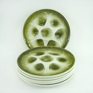 Puede incluir: Un conjunto de seis platos de cerámica verde y blanca para ostras. Cada plato tiene seis compartimentos en forma de concha alrededor de un pozo central. Los platos tienen un acabado brillante y están apilados sobre un fondo blanco.