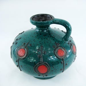 Peut inclure: Un vase en céramique bleu-vert avec un glaçage moucheté et des accents rouges et noirs. Le vase a un corps rond et une seule poignée.