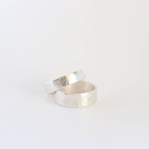 Può includere: Due anelli d'argento con una texture martellata sono impilati. Gli anelli hanno un design semplice e minimalista. L'anello superiore è più sottile di quello inferiore. Gli anelli sono probabilmente in argento sterling o metallo simile.
